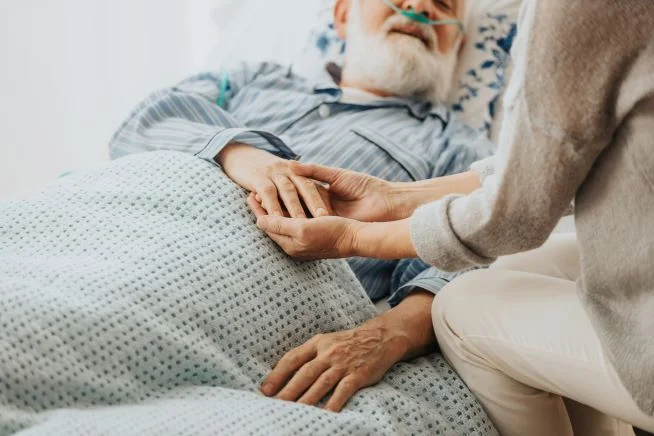 Ein älterer, kranker Herr mit weißem Bart und geschlossenen Augen liegt in einem Bett. Eine zweite Person, von der man nur die Hände und den Pullover sieht,  steht am Bett und hält die Hände des alten Mannes. 